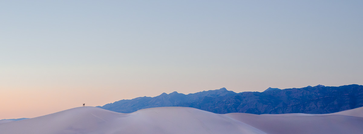 Two figures traverse luminous white dunes beneath a pastel twilight sky.