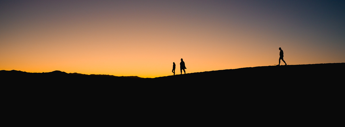 Three silhouettes walk a dune ridge against a blazing desert sunset.