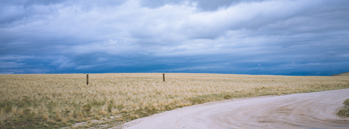 A dirt road curves past golden grasslands under gathering storm clouds.