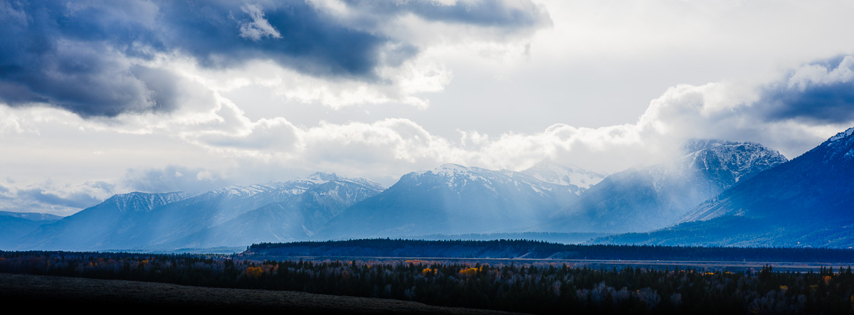 Light breaks through storm clouds over snow-dusted mountains and autumn forests.