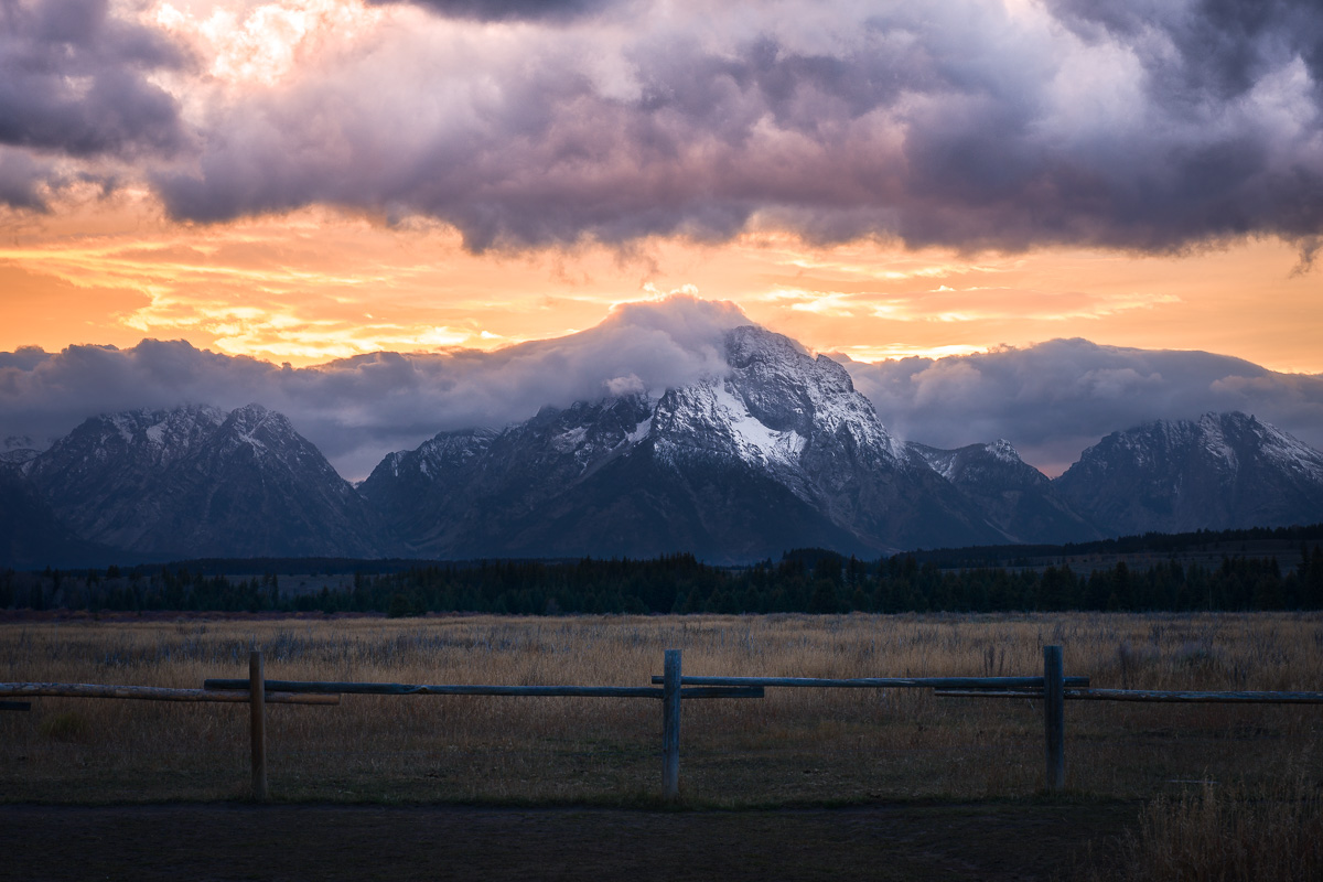 Storm clouds crown the Grand Tetons in fire as a wooden fence frames the valley.