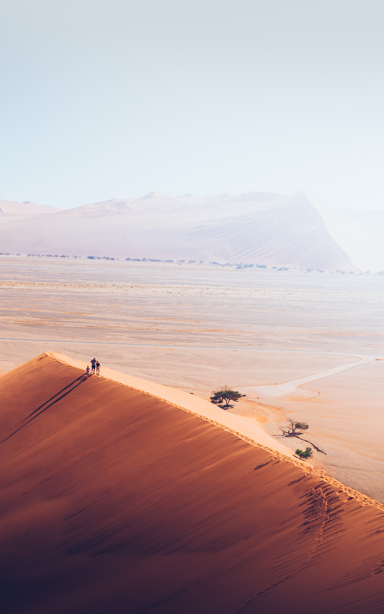 Tiny figures ascend a dune ridge dwarfed by hazy desert beyond.